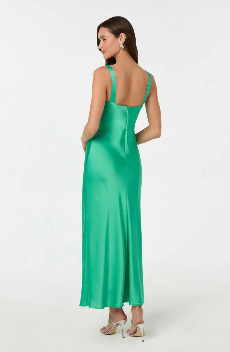 A woman is wearing a floor-length, sleeveless gown in a vibrant, shiny green satin fabric. The dress has wide shoulder straps and a fitted bodice, flowing smoothly down to the hem. She is also wearing white high-heeled shoes with thin straps and large, sparkling earrings.