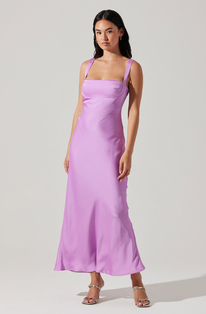 A woman is wearing a sleeveless, full-length lavender satin dress with wide straps and a fitted bodice. The fabric has a shiny, smooth texture, and the dress flows down to her ankles. She is also wearing silver strappy high-heeled sandals.