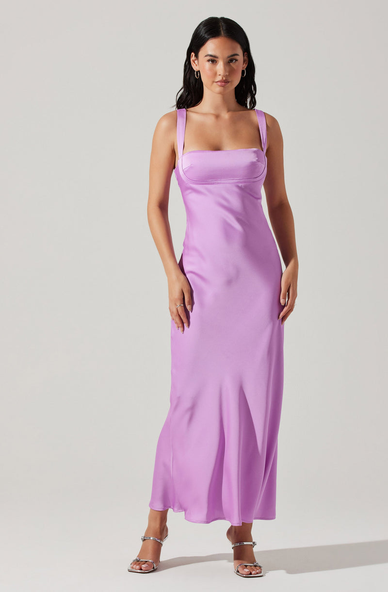A woman is wearing a sleeveless, lavender-colored dress with thick shoulder straps. The bodice is fitted with a horizontal seam across the chest, and the skirt flows down to the floor with a subtle slight flare near the hem. She is also wearing open-toe high-heeled sandals with ankle straps.