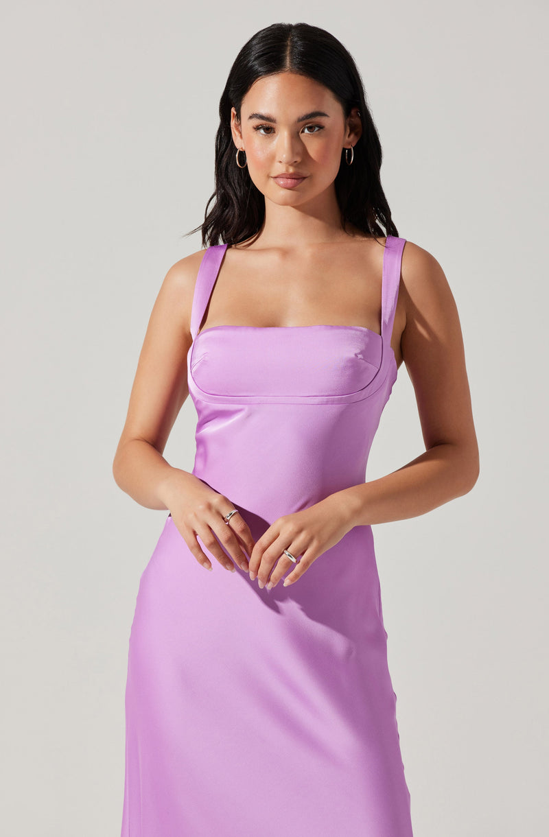 A woman is wearing a sleeveless, lavender-colored dress with wide shoulder straps. The dress has a fitted bodice with a horizontal seam across the chest and a smooth, flowing skirt.