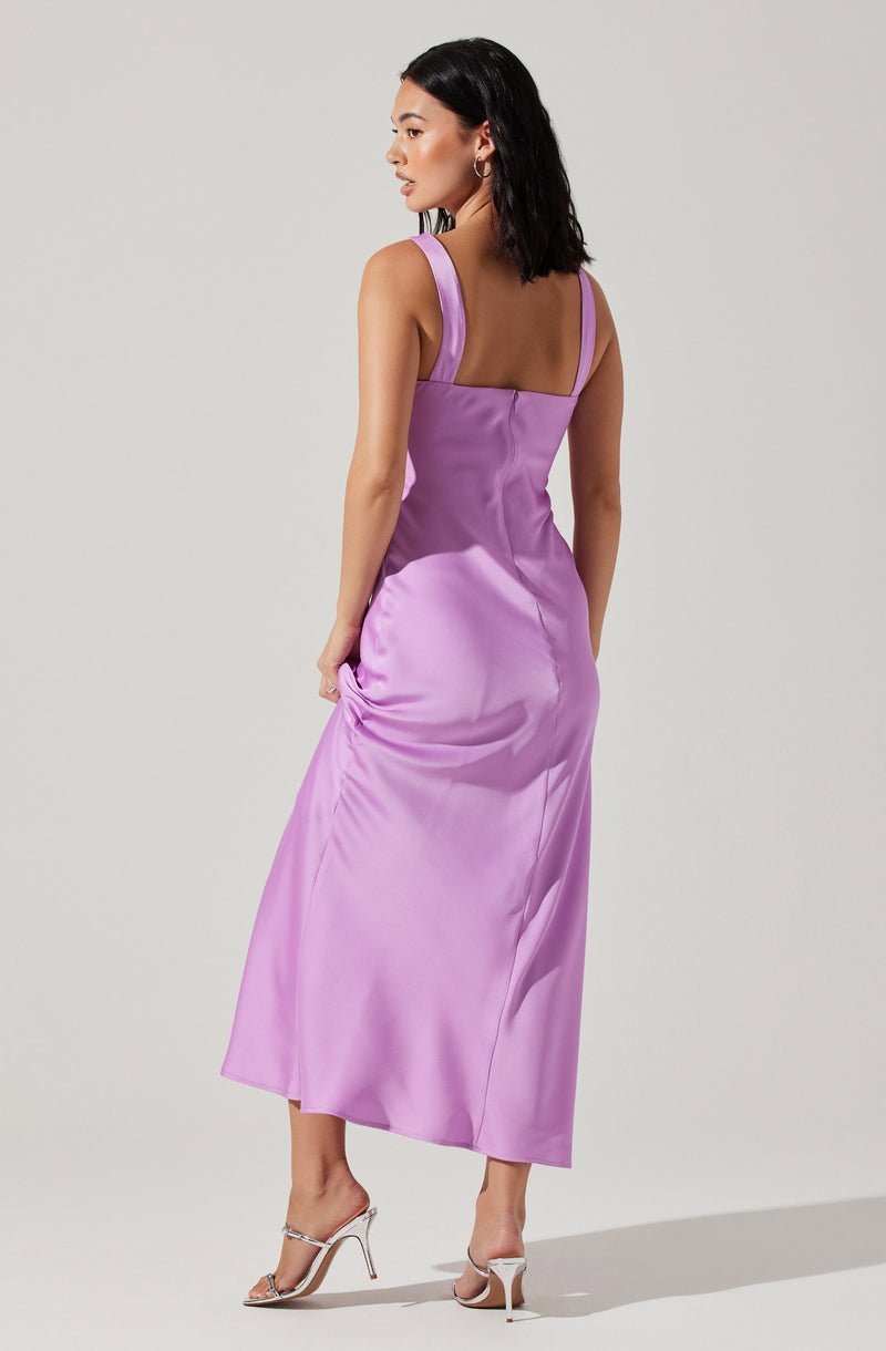 A woman is wearing a sleeveless, lavender satin dress with thick shoulder straps and a straight neckline. The dress is fitted at the waist and flows down to a midi length, ending above her ankles. She is wearing white high-heeled sandals.