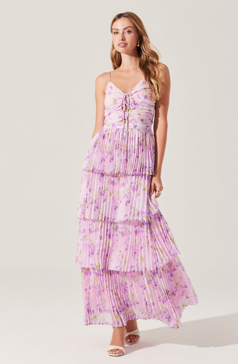 A woman is wearing a long, tiered maxi dress with thin spaghetti straps. The dress features a light pink and purple floral pattern with gold accents, and each tier is pleated, creating a flowing, layered look. The top has a lace-up detail at the neckline.