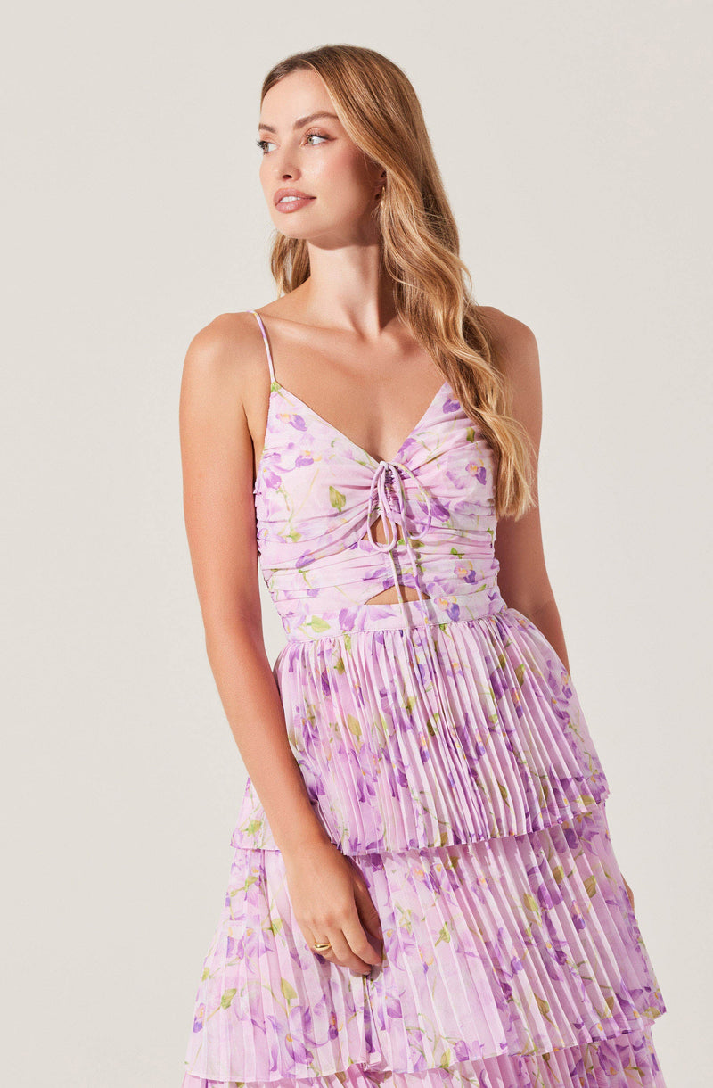 A woman is wearing a sleeveless dress with thin spaghetti straps. The dress features a fitted bodice with a tie detail at the front, and the fabric has a pastel purple base with a floral pattern incorporating shades of green and lavender. The skirt has multiple layers of pleated fabric, creating a tiered, ruffled effect.