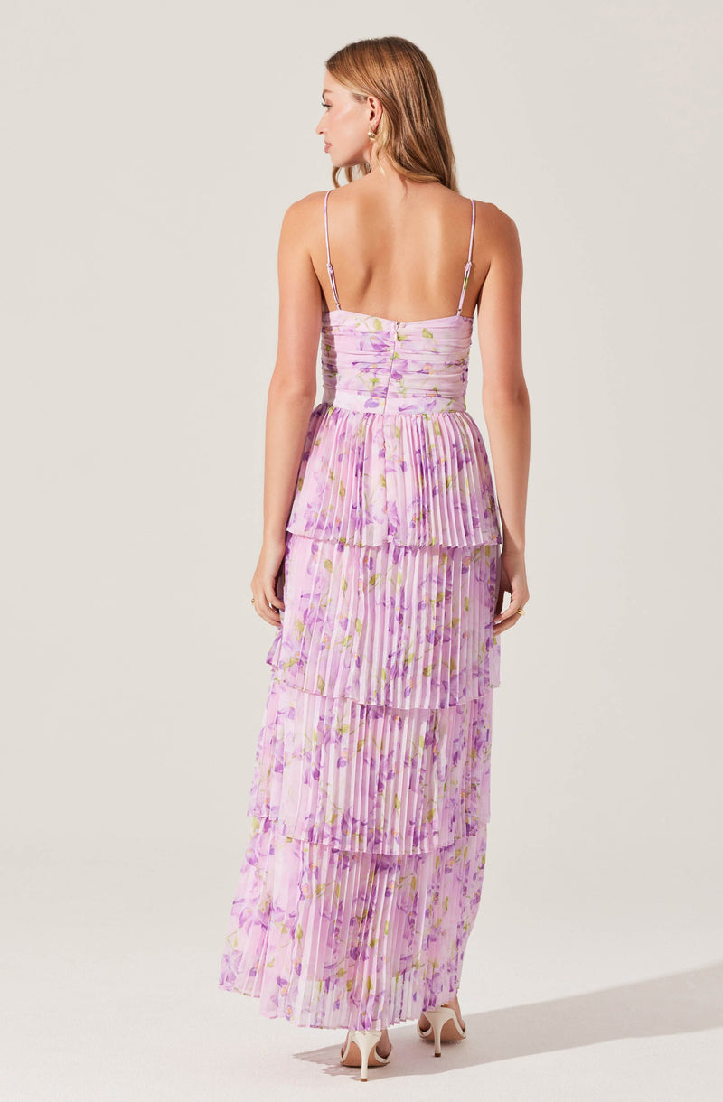 A woman is wearing a sleeveless, floor-length dress with thin spaghetti straps. The dress features a pastel pink and purple floral pattern with vertical pleats throughout. The bodice is fitted with ruched detailing, and the skirt has tiered, ruffled layers that create a flowing silhouette. She is also wearing nude high-heeled shoes.