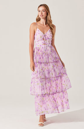 A woman is wearing a long, tiered maxi dress with thin spaghetti straps. The dress features a purple and pink floral pattern with vertical stripes. The upper part has a ruched design with a small tie in the center of the bust. The dress is made of a lightweight, flowy fabric and has four horizontal tiers, each with a slightly ruffled seam, creating a layered and textured look. She is also wearing a pair of white open-toe sandals.