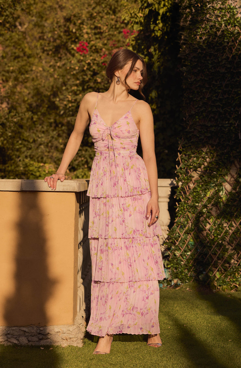 A woman is wearing a pink, tiered, floral maxi dress with a fitted bodice and thin spaghetti straps. The dress features a subtle tie detail at the front of the neckline and has a flowy, layered skirt with a delicate, colorful flower pattern. She accessorizes with large hoop earrings and a ring on her right hand.