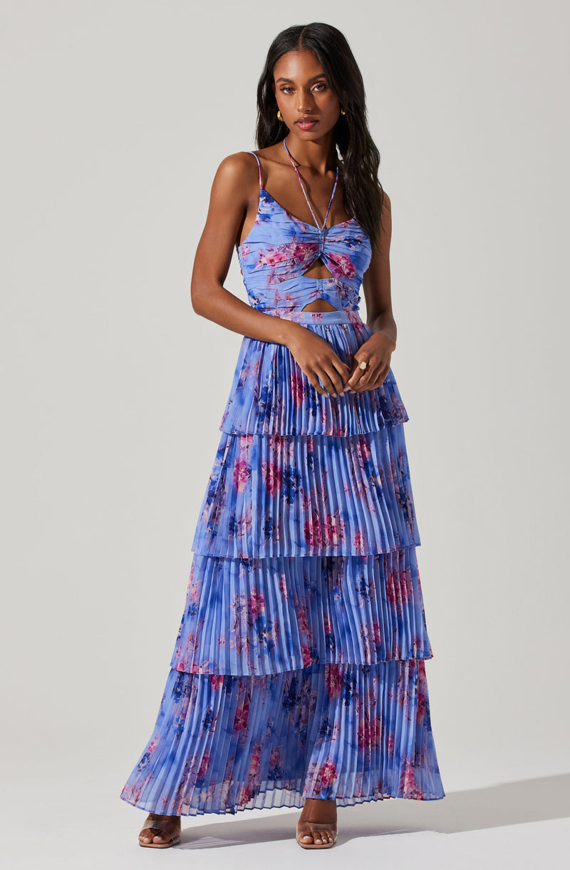 A woman is wearing a matching two-piece outfit with a sleeveless, spaghetti strap crop top that features a knot detail in the center and cut-out sections on the front. The fabric has a vibrant blue background with a pink and purple floral pattern. The matching high-waisted, tiered skirt is pleated with multiple layers, each with the same floral pattern, creating a flowing, tiered design. She is also wearing a long necklace and open-toed heels.
