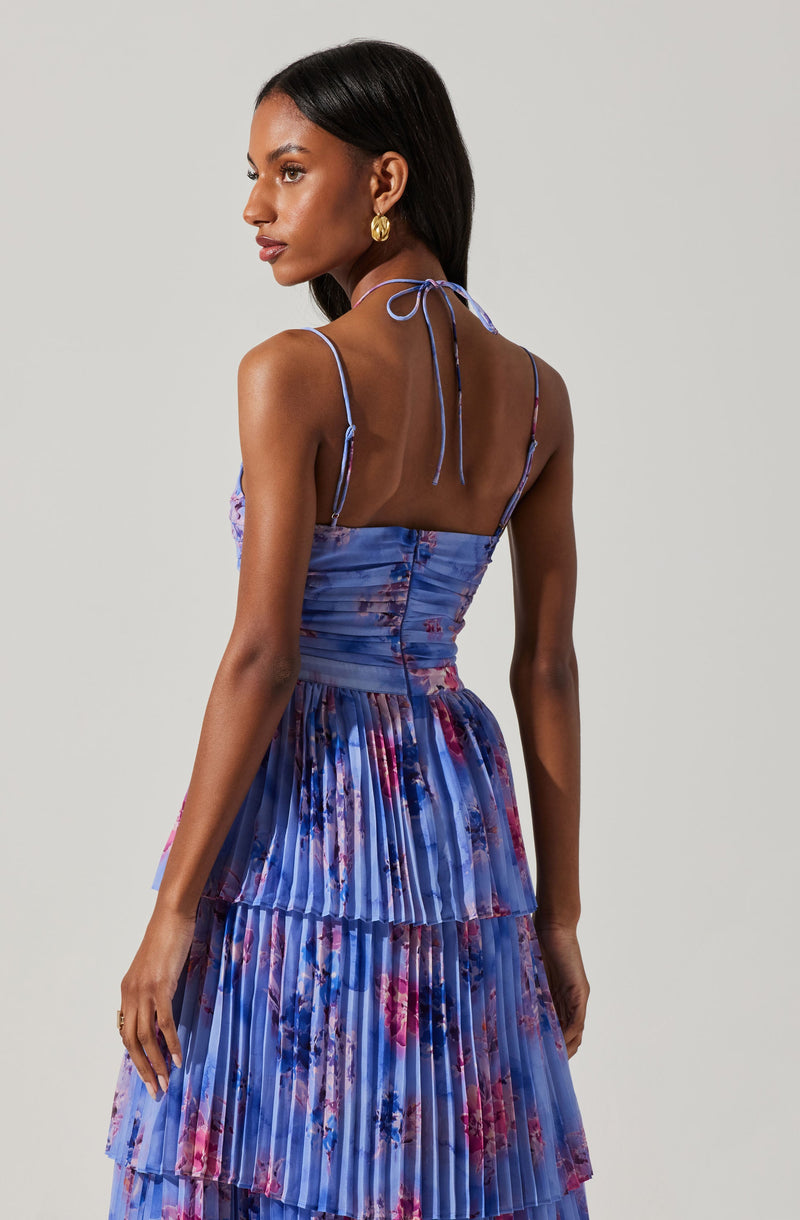 A woman is wearing a sleeveless dress with adjustable spaghetti straps, tied at the back of her neck. The dress has a fitted bodice and a tiered, pleated skirt. The fabric features a blue base color with a floral pattern in shades of pink and purple.