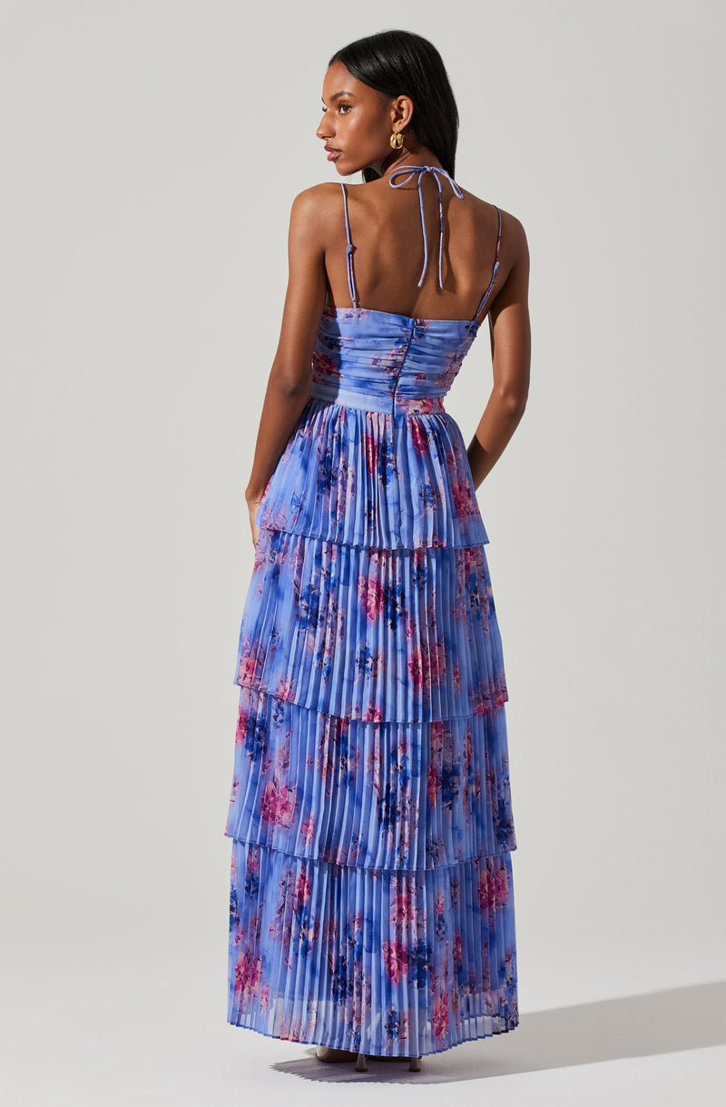 A woman is wearing a sleeveless, floor-length dress with thin, adjustable spaghetti straps tied in bows at the shoulders. The dress features a fitted bodice with a ruched design and a flowy, tiered skirt, all made from a pleated fabric with a floral pattern in shades of blue, pink, purple, and red.