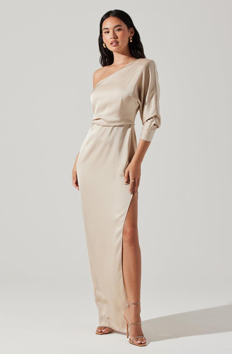 A woman is wearing a long, elegant, cream-colored satin dress with an asymmetrical, one-shoulder neckline. The dress features a single sleeve that is gathered at the wrist, a waistline that cinches slightly, and a high slit on the side revealing her leg. She is also wearing high heels with ankle straps and gold earrings.