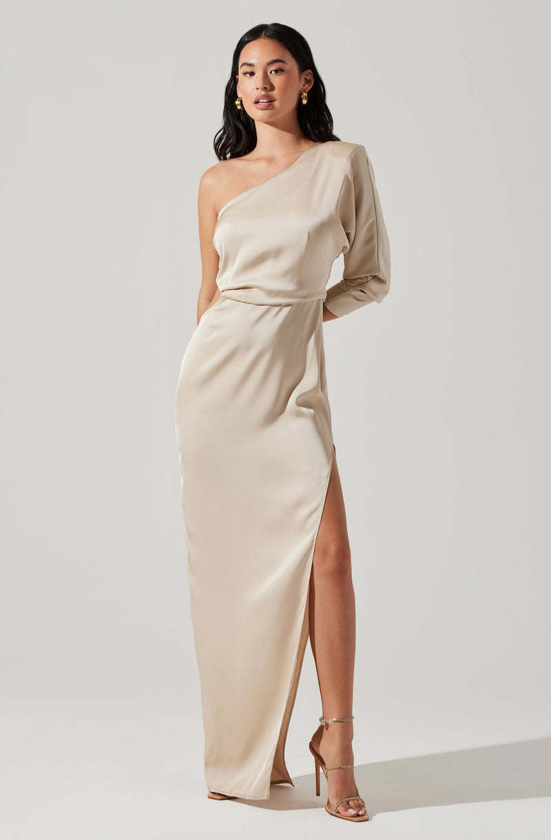 A woman is wearing a cream-colored, one-shoulder evening gown. The dress features a single long sleeve on the left arm, while the right shoulder and arm are bare. The gown has a high slit on the left side, revealing her leg and a strappy high-heeled sandal.