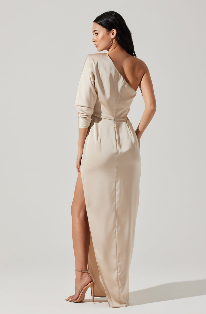 A woman is wearing a long, elegant cream-colored satin dress with a high slit on one side, revealing her leg. The dress features an asymmetrical off-the-shoulder design, with one side having a long sleeve while the other shoulder is bare. She is also wearing nude high-heeled sandals.