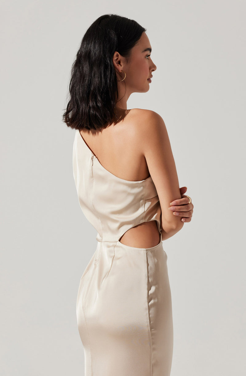 A woman is wearing a shiny, satin-like cream-colored dress with a one-shoulder design. The dress features a cutout detail at the waist, revealing a small section of her midriff. She has shoulder-length dark hair, wears hoop earrings, and is standing in profile with her arms crossed.