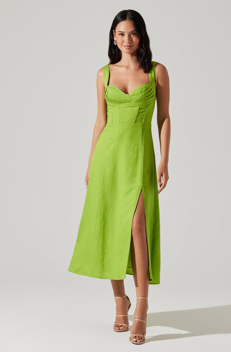 A woman is wearing a sleeveless, bright green maxi dress with wide shoulder straps. The dress features a fitted bodice with ruched detailing across the bust and a high slit on the left side, exposing her leg. She is also wearing nude-colored high-heeled sandals.