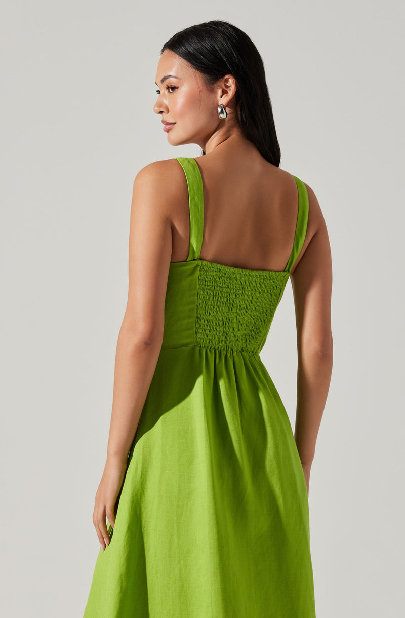 A woman is wearing a bright green sleeveless dress with wide shoulder straps. The dress features a fitted, smocked bodice with a textured, shirred design, and the skirt is gathered at the waist, flowing outward. The fabric appears to be shiny, possibly satin or similar material. The woman has long, dark hair and is accessorized with large, silver hoop earrings.