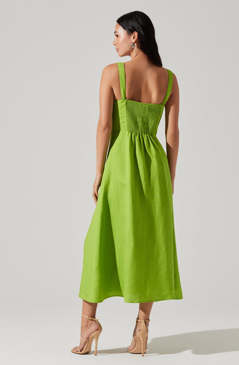 A woman is wearing a bright green sleeveless dress with wide shoulder straps. The dress features a gathered, smocked bodice and flows down to a full-length skirt. She is also wearing nude high-heeled sandals.