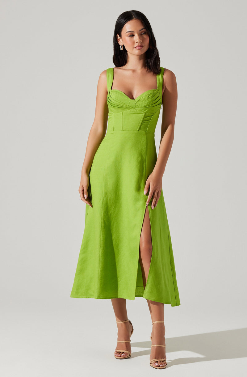 A woman is wearing a sleeveless, lime green dress with broad shoulder straps and a fitted bodice featuring ruching details. The dress has a high slit on one side, revealing her leg. She is also wearing beige high-heeled sandals with ankle straps.