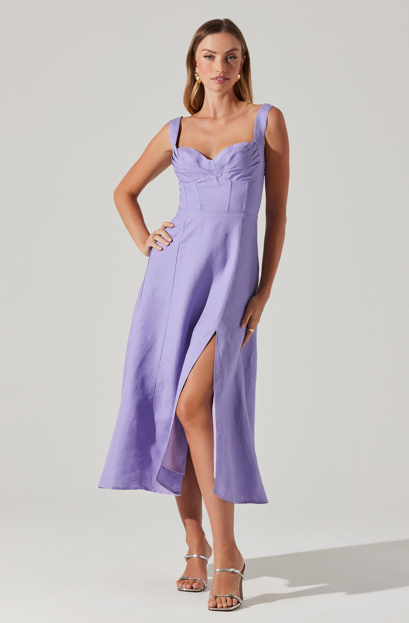 A woman is wearing a sleeveless, lavender-colored dress with wide shoulder straps, a fitted bodice with pleated detailing, and a high slit on the left side that reveals her leg. She pairs the dress with open-toe, ankle-strap heels and gold earrings.