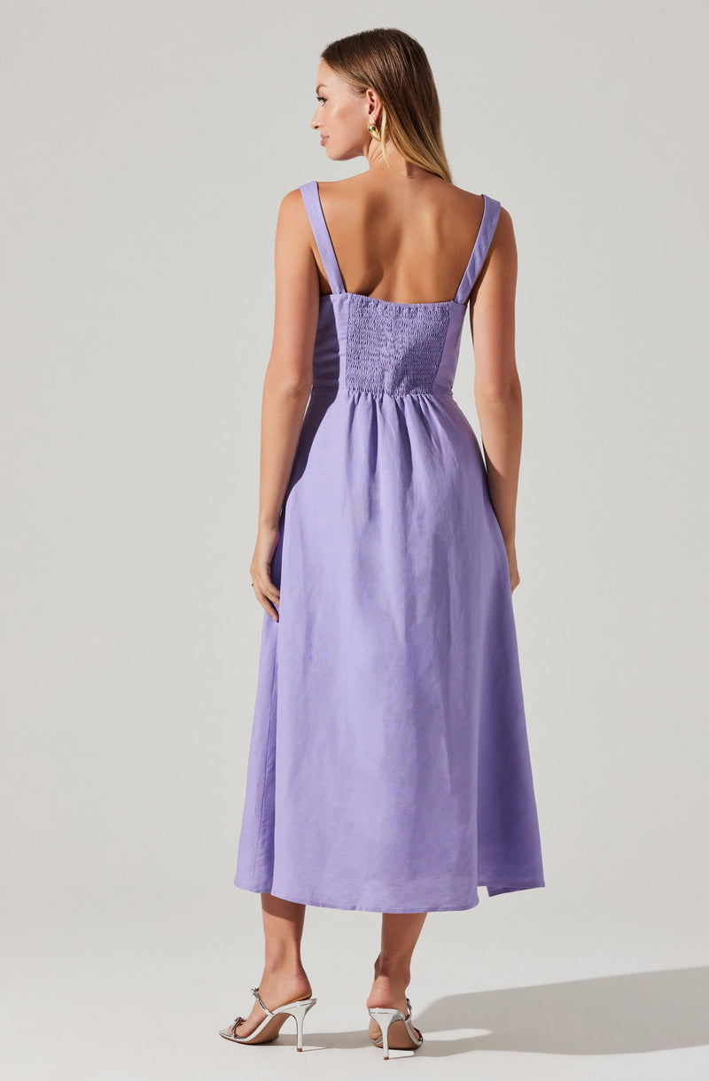 A woman is wearing a sleeveless, lavender-colored dress with wide shoulder straps. The dress features a fitted, smocked bodice and a full, flowing skirt that reaches mid-calf. She is also wearing a pair of open-toed, high-heeled sandals.