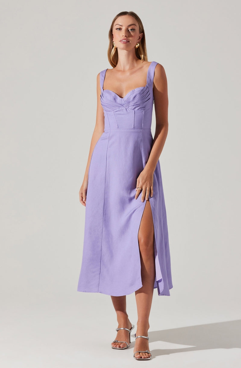 A woman is wearing a sleeveless, lavender-colored dress with a fitted bodice featuring pleated details and wide shoulder straps. The dress has a high slit on the left side, revealing her leg. She pairs the dress with strappy, silver high-heeled sandals.