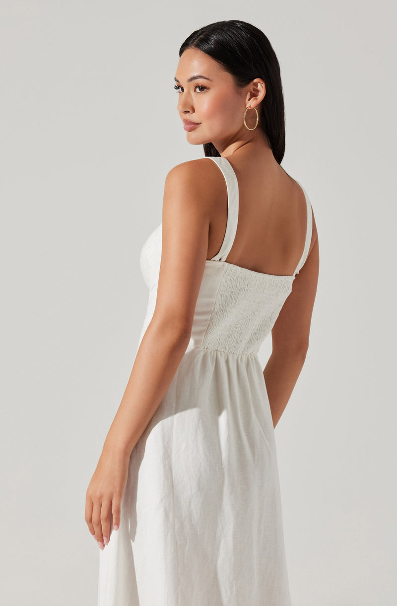 A woman is wearing a sleeveless, white, full-length dress with wide shoulder straps. The top section of the dress features a shirred, fitted bodice, and the skirt flows loosely from the waist. She accessorizes with large, gold hoop earrings and has straight, dark hair.