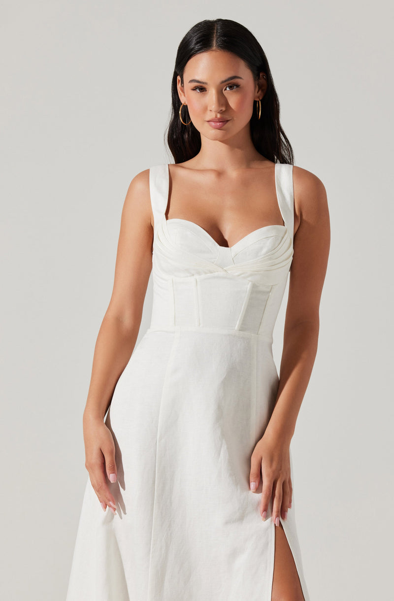 A woman is wearing a sleeveless white dress with wide straps and a sweetheart neckline. The bodice features pleated detailing that creates a fitted waist, and the skirt has a high slit on the right side. She accessorizes with gold hoop earrings.