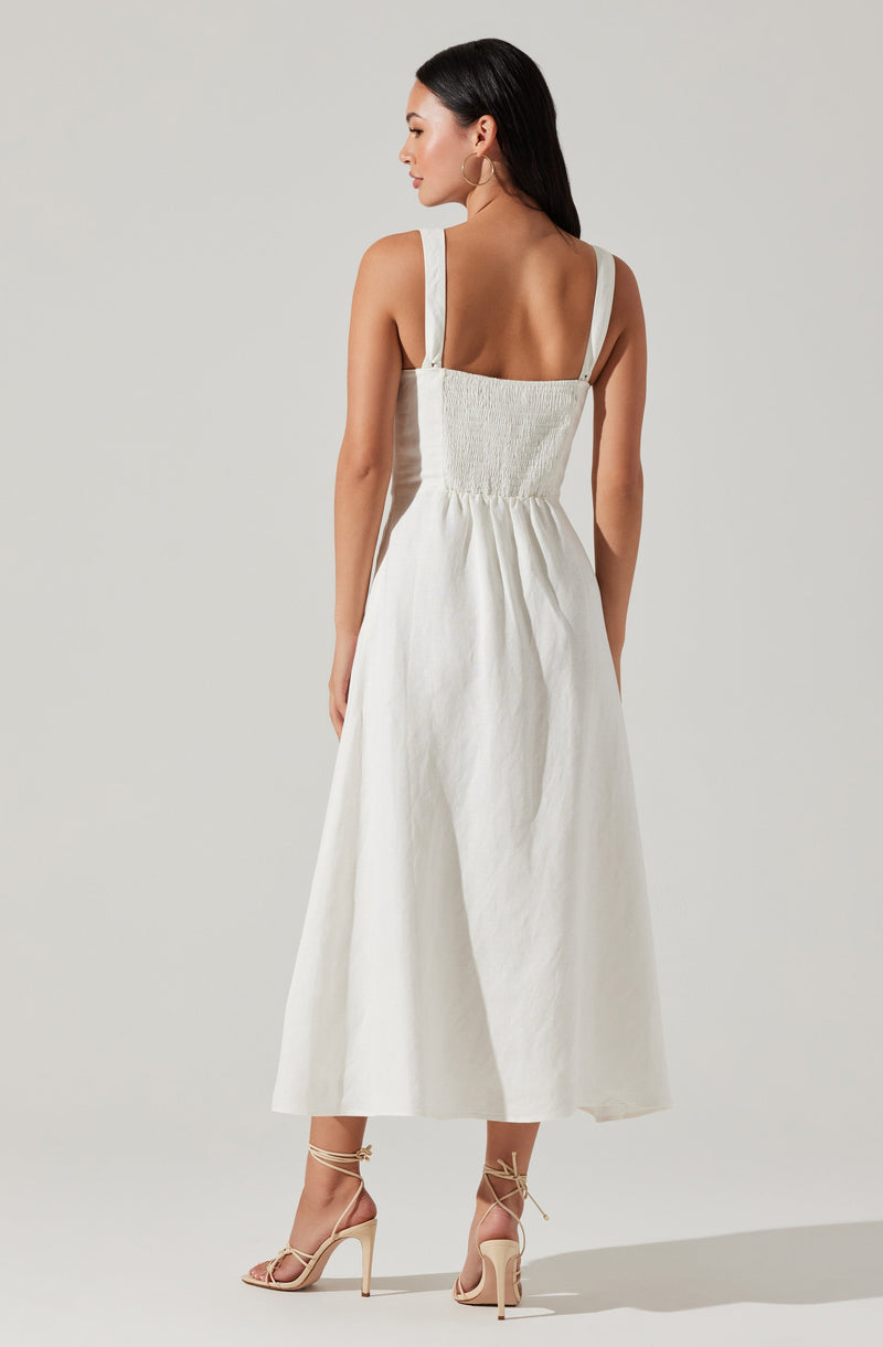 A woman is wearing a sleeveless, white, ankle-length dress with wide shoulder straps and a smocked bodice. She is also wearing beige high-heeled sandals with ankle straps.
