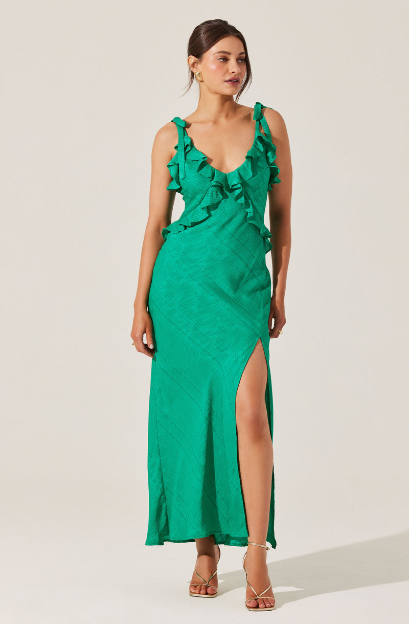 A woman is wearing a sleeveless, emerald green dress with ruffle details along the neckline and straps. The dress features a thigh-high slit on the front left side, revealing her leg. She is also wearing strappy high-heeled sandals.