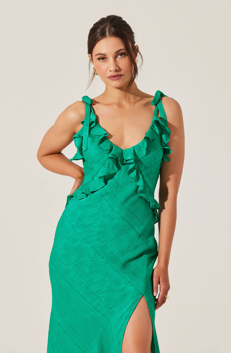 A woman is wearing a sleeveless, green dress with ruffled shoulder straps and a ruffle detail running diagonally across the bodice. The dress has a subtle checkered pattern and features a high slit on the left side.