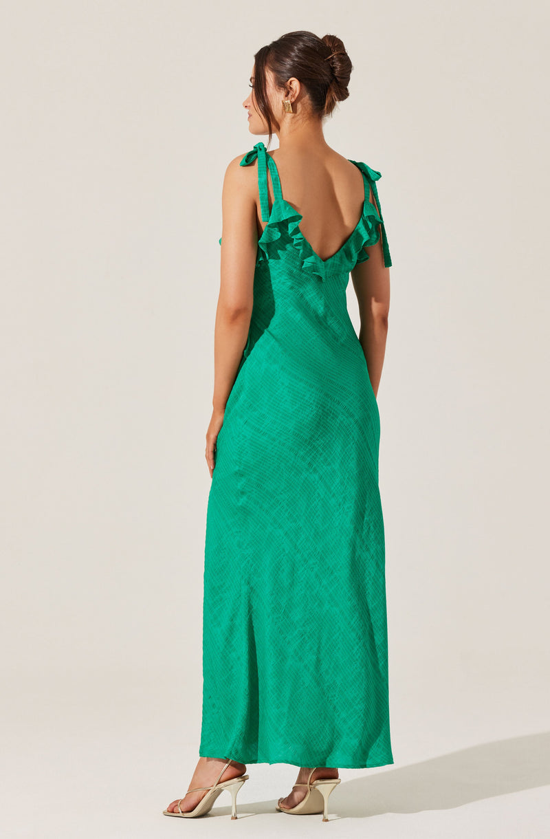 A woman is wearing a sleeveless, floor-length dress in a bright green color. The dress features shoulder tie straps with bow details and ruffle accents along the neckline and shoulder straps. The fabric has a textured, woven pattern, and the dress has a deep V-back. She is also wearing beige high-heeled sandals.