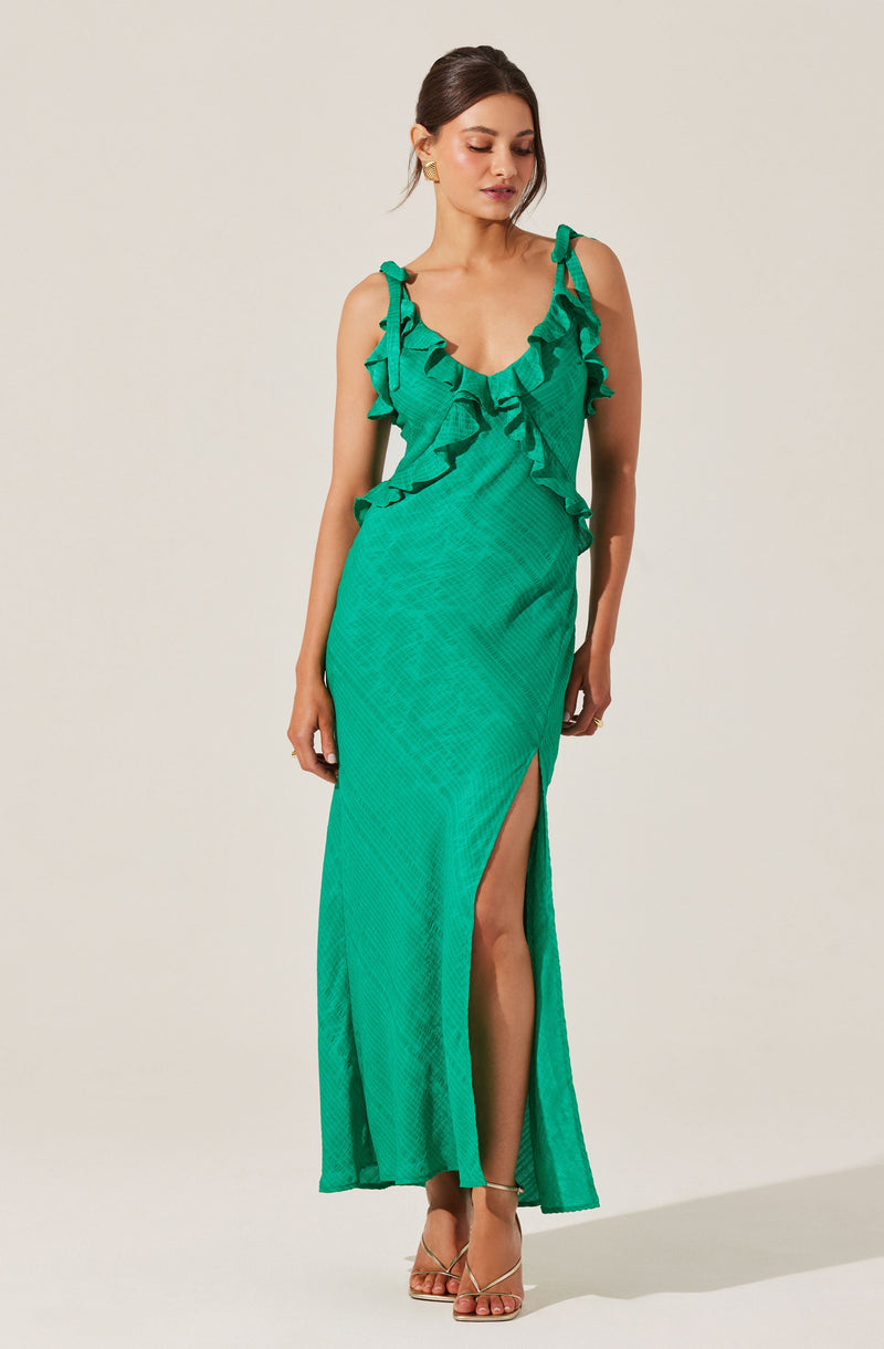A woman is wearing a long, sleeveless green dress with ruffle details along the neckline and shoulders. The dress has a fitted waist and features a high slit on one side, revealing her leg. She is also wearing strappy high-heeled sandals.