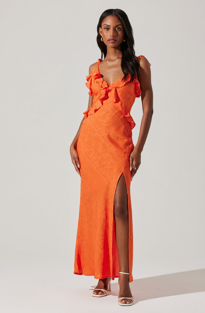 A woman is wearing a sleeveless, full-length orange dress with ruffle details along the neckline and shoulders. The dress features a thigh-high slit on one side, revealing her leg. She is also wearing open-toe, high-heeled sandals with ankle straps.