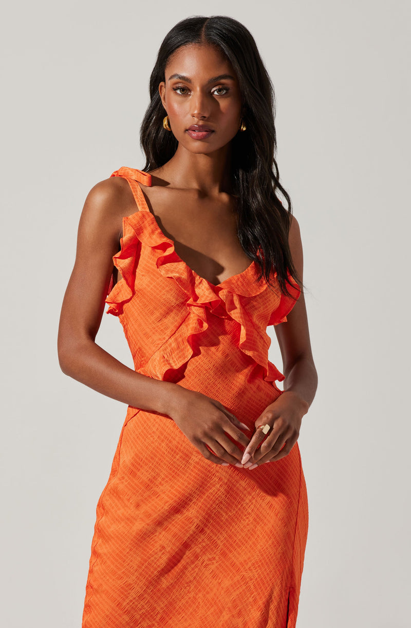 A woman is wearing a sleeveless, orange dress with ruffle details along the neckline and straps. The fabric has a subtle textured pattern, and the dress fits snugly around her waist. She accessorizes with gold hoop earrings and a large ring on her right hand.