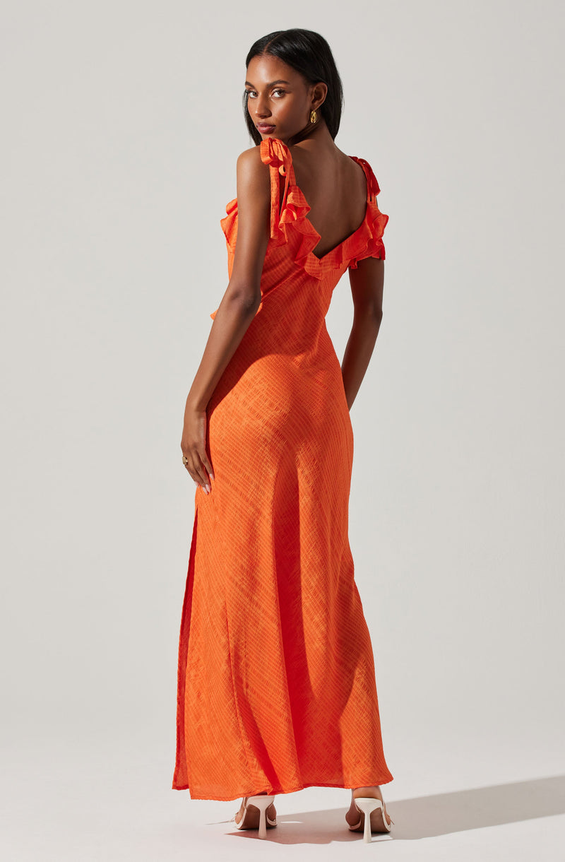 A woman is wearing a long, orange dress with ruffled shoulder straps. The dress has a textured, checkered pattern and features a deep V-back. She is also wearing white high-heeled shoes.