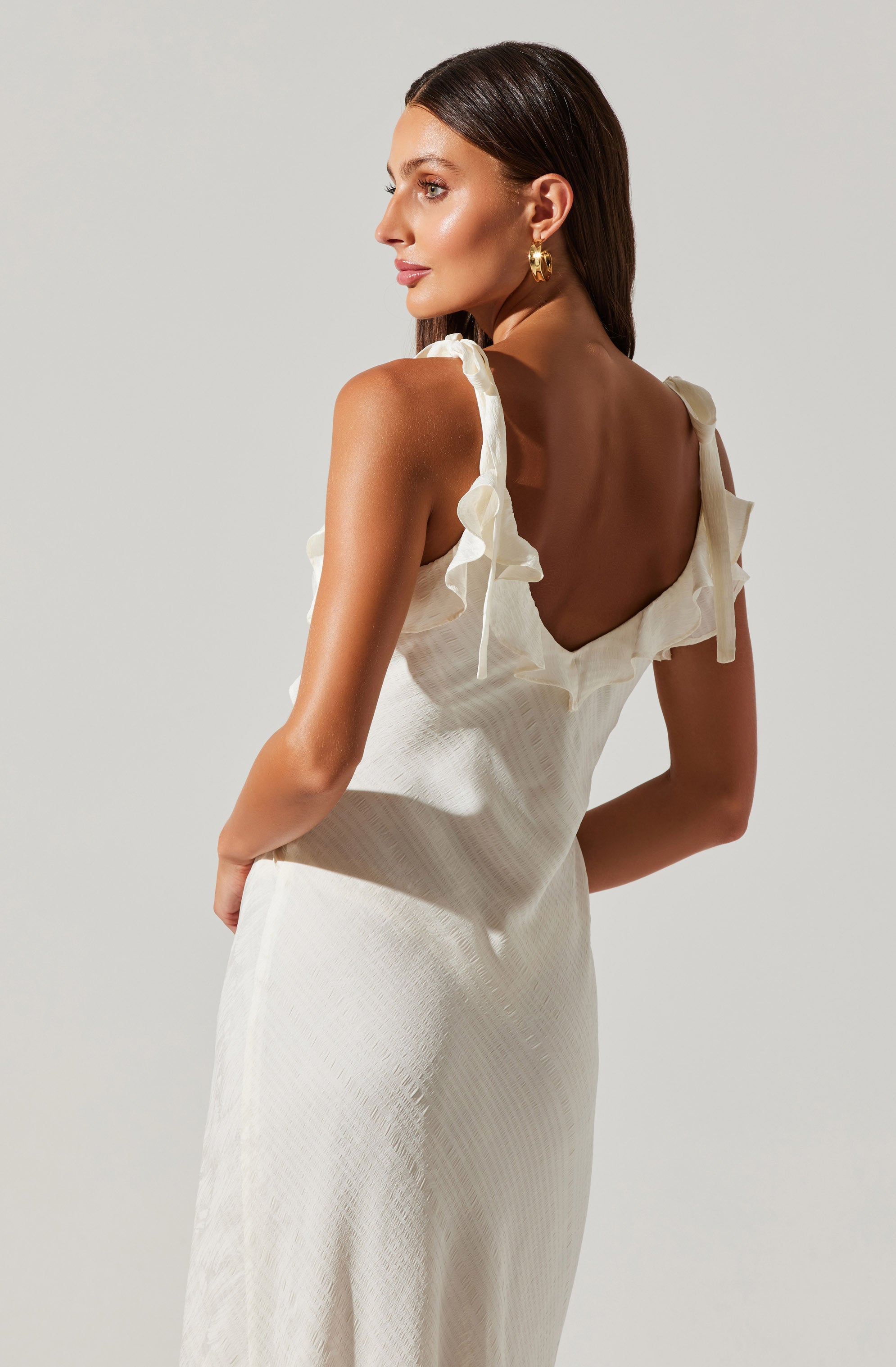 A woman is wearing a sleeveless, cream-colored dress with ruffled shoulder straps and a deep V-back. The fabric appears to be textured with a subtle pattern.