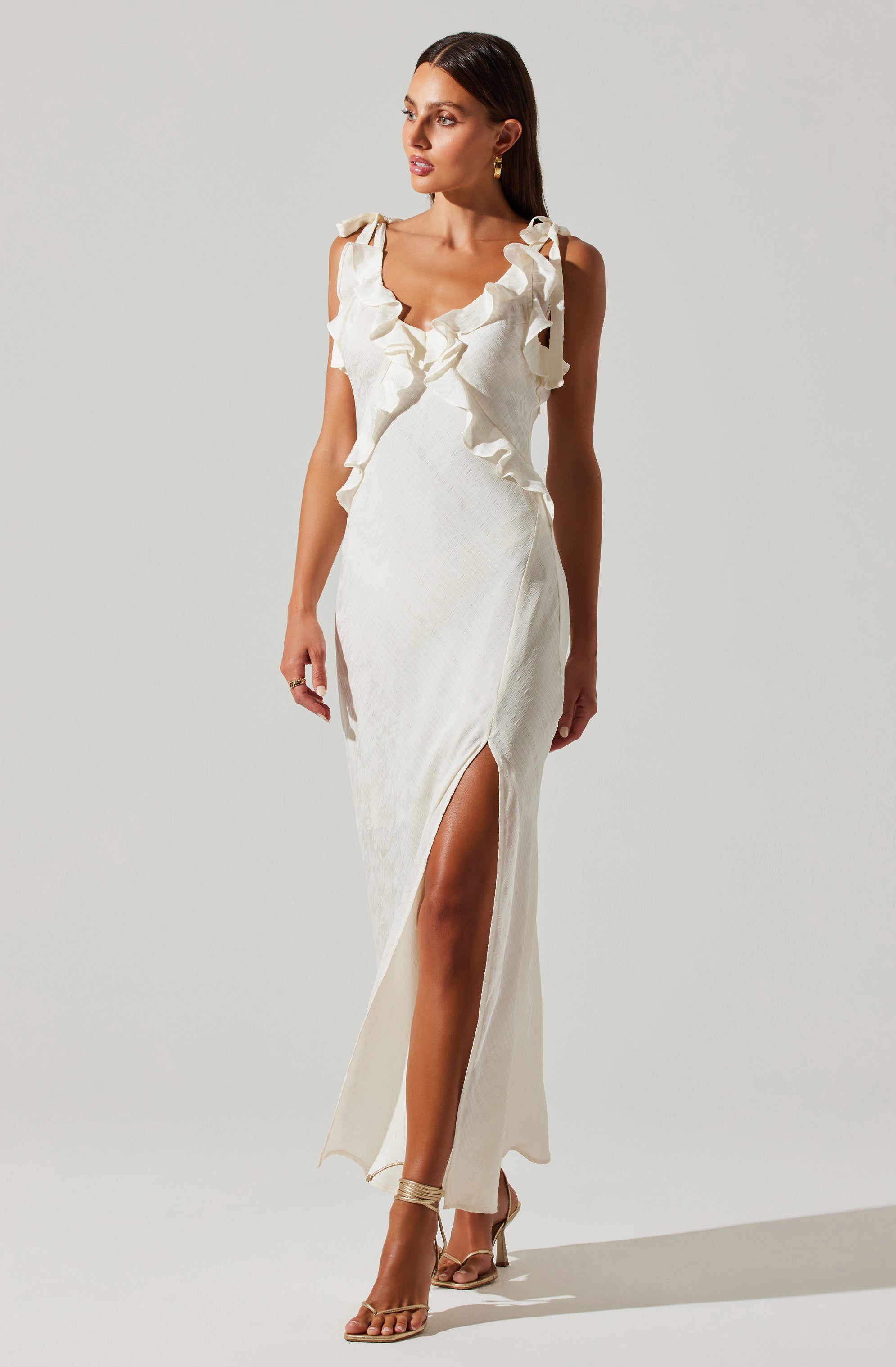 A woman is wearing a long, elegant white dress with ruffled detailing around the shoulders and neckline. The dress features a high slit on one side, revealing her leg. She is also wearing strappy high-heeled sandals.