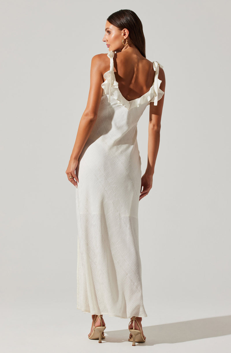 A woman is wearing a long, sleeveless white dress with ruffled straps that tie at the shoulders. The dress has a deep V-neckline with a ruffled trim along the edges. She is also wearing beige high-heeled sandals with ankle straps.