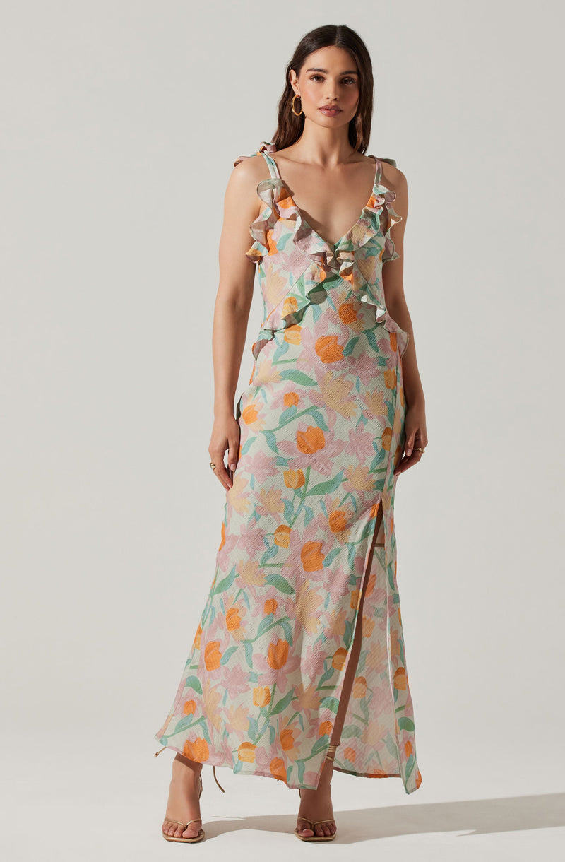 A woman is wearing a long, flowy dress with a colorful floral pattern featuring shades of pink, orange, green, and beige. The dress has ruffled shoulder straps and a plunging V-neckline. It also has a high slit on one side, revealing part of her leg. She is accessorized with large, circular hoop earrings and neutral-colored high-heeled sandals.