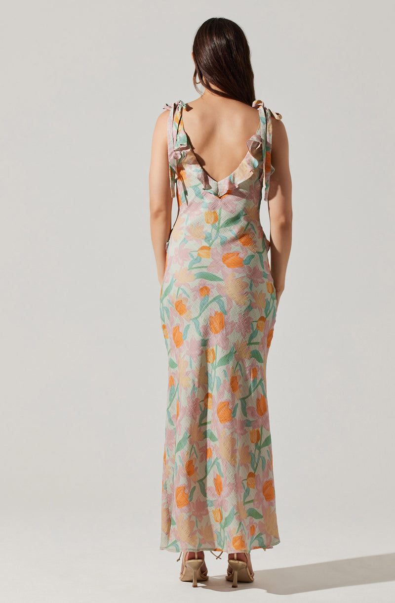 The dress is a sleeveless, full-length gown with a V-shaped back, featuring shoulder ties. The fabric has a colorful, abstract floral pattern in shades of orange, pink, green, and beige.