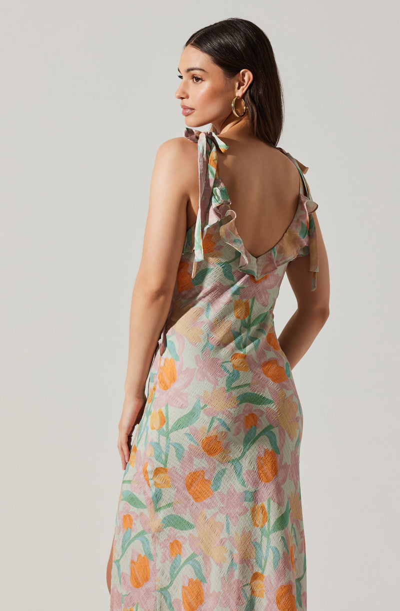 A woman is wearing a sleeveless dress with shoulder straps tied into bows. The dress features a colorful, abstract floral pattern with shades of orange, pink, green, and beige. The fabric appears lightweight and textured, with ruffled details along the shoulder straps.