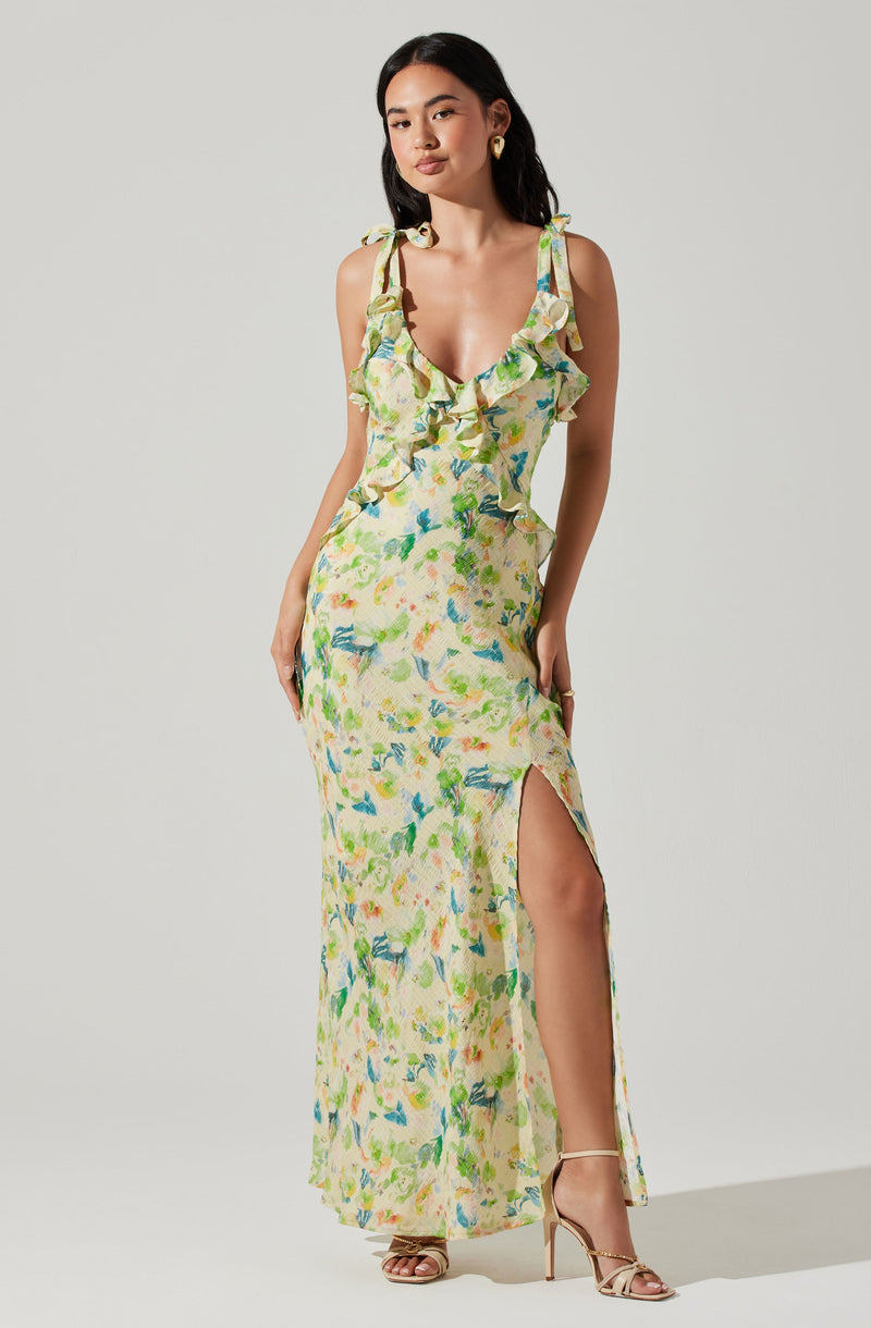 A woman is wearing a long, sleeveless dress with shoulder ties and a ruffled neckline. The dress features a colorful, abstract floral pattern in shades of yellow, green, blue, and pink. It has a high slit on one side, revealing her leg. She is also wearing gold hoop earrings and beige high-heeled sandals.