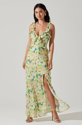 The dress is a sleeveless, ankle-length summer gown with shoulder straps tied into bows. It features a deep V neckline with ruffles along the front. The fabric has a colorful, abstract floral pattern in shades of green, blue, yellow, and orange. The dress includes a high slit on one side, revealing the leg, and the material appears lightweight and flowy.