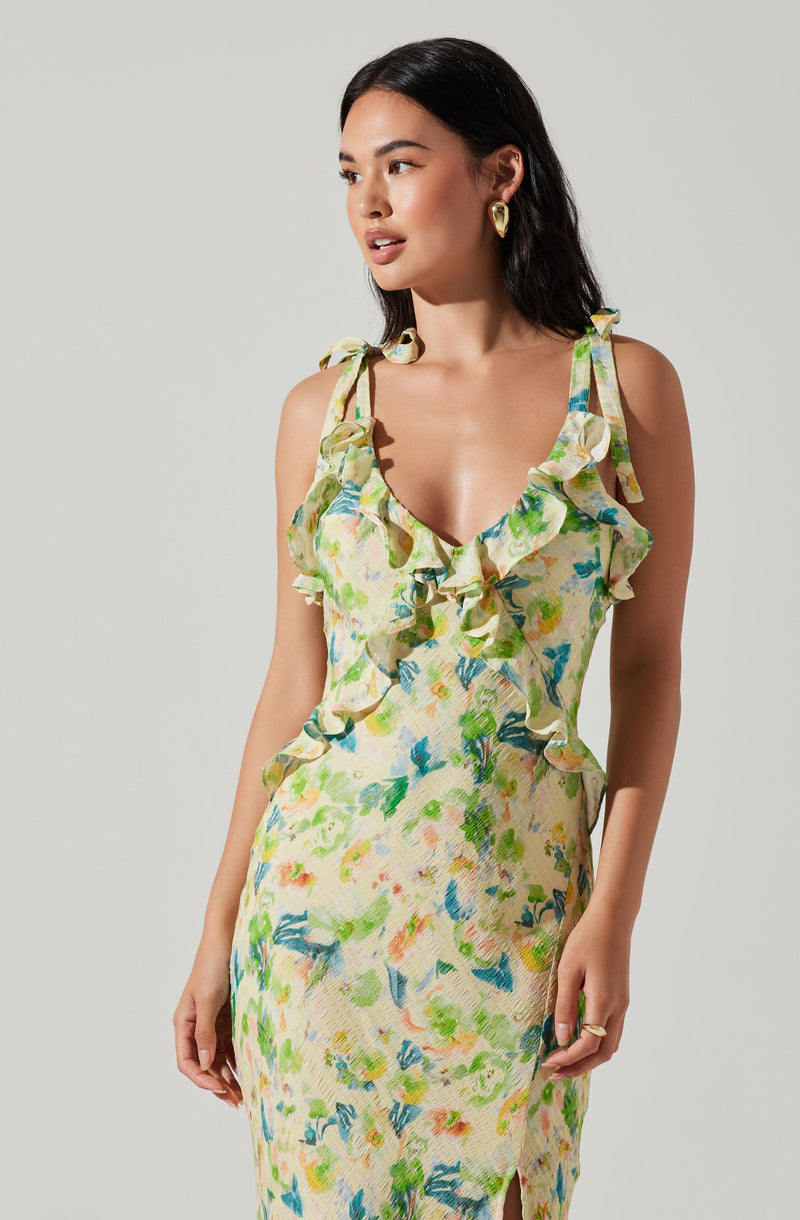 A woman is wearing a sleeveless dress with shoulder straps tied into bows. The dress features a colorful, abstract floral pattern with shades of green, blue, yellow, and orange. It has ruffled detailing along the neckline and shoulder straps, creating a feminine and playful look.