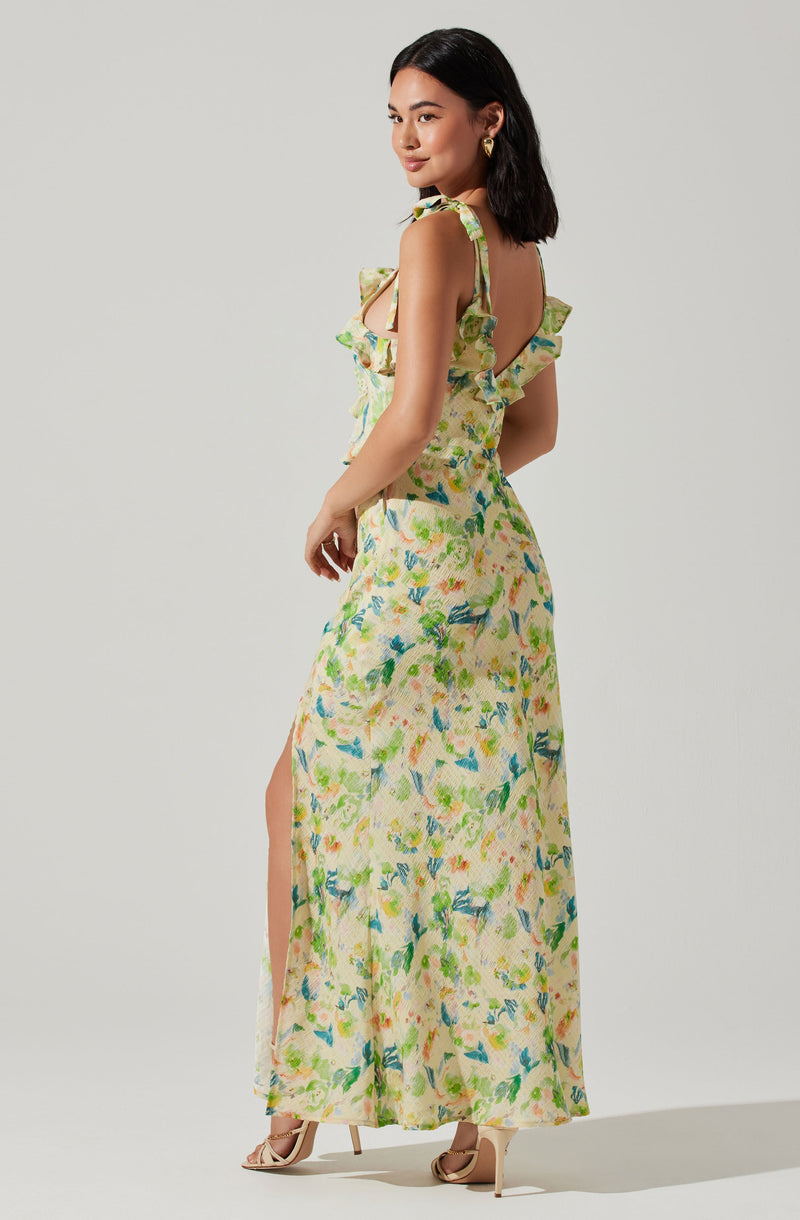 A woman is wearing a sleeveless, ankle-length dress with a light, flowy fabric featuring a colorful, abstract floral pattern in shades of green, yellow, blue, and pink. The dress has ruffled shoulder straps and a cut-out design at the back. It also has a slit on one side, revealing her leg. She is wearing nude-colored high heels.