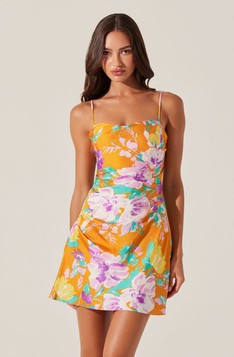 A young woman is wearing a sleeveless, floral summer dress with thin spaghetti straps. The dress features a vibrant pattern of large flowers in shades of purple, pink, yellow, and teal on a bright orange background.