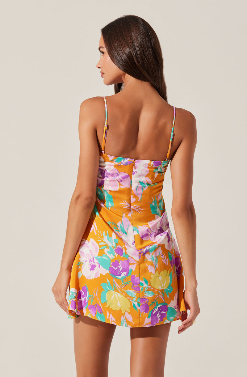 A woman is wearing a sleeveless, fitted dress with thin spaghetti straps. The dress features a vibrant floral pattern with large blossoms in shades of purple, pink, yellow, green, and white on an orange background.