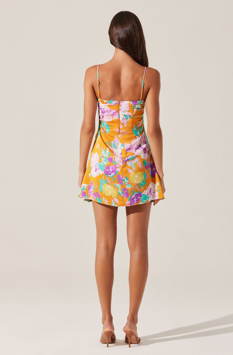 A woman is wearing a sleeveless, above-the-knee dress with thin spaghetti straps. The dress features a vibrant floral pattern with shades of orange, purple, pink, green, and blue. The fabric appears silky with a slight sheen, and the dress has a fitted bodice that flares out into a short skirt.