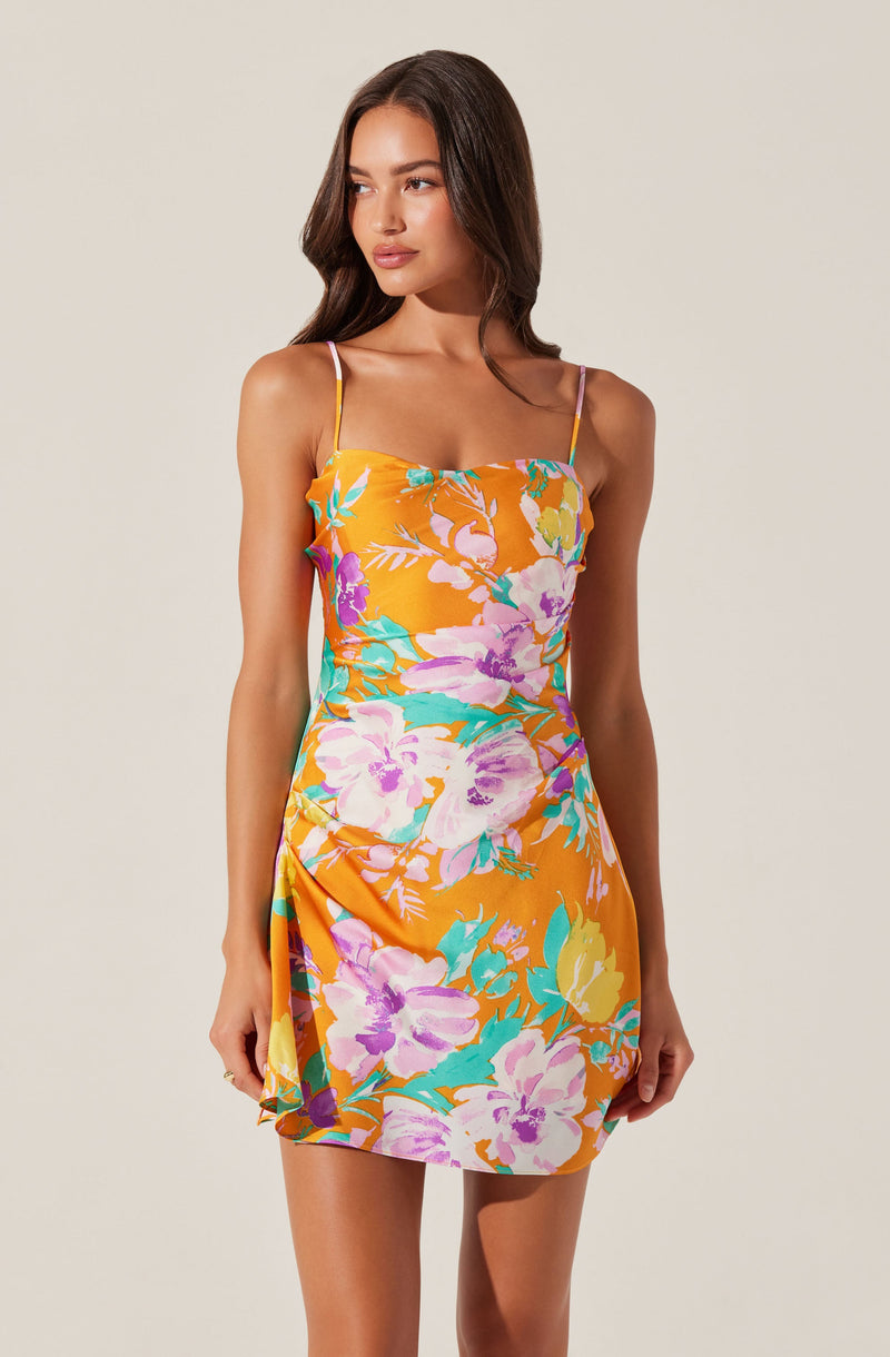 A woman is wearing a sleeveless dress with thin spaghetti straps. The dress features a bright orange background with a large floral pattern in shades of purple, pink, yellow, and teal. The dress has a fitted bodice and a slightly gathered detail on one side, creating a subtle drape effect.