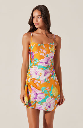 A woman is wearing a sleeveless dress with thin spaghetti straps. The dress features a bright orange background with a large floral pattern in shades of purple, pink, yellow, and teal. The dress has a fitted bodice and a slightly gathered detail on one side, creating a subtle drape effect.