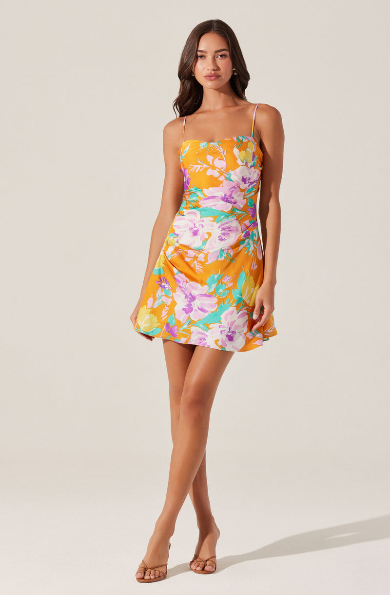 A young woman is wearing a sleeveless, floral print summer dress with thin spaghetti straps. The dress features a bright orange background with large, colorful flowers in shades of pink, purple, yellow, and teal. The hem of the dress is above the knee, and she is wearing nude-colored sandals.